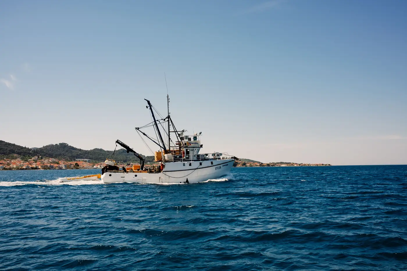 A fishing trawler with visible gear and rigging sails across open water near a coastal town.