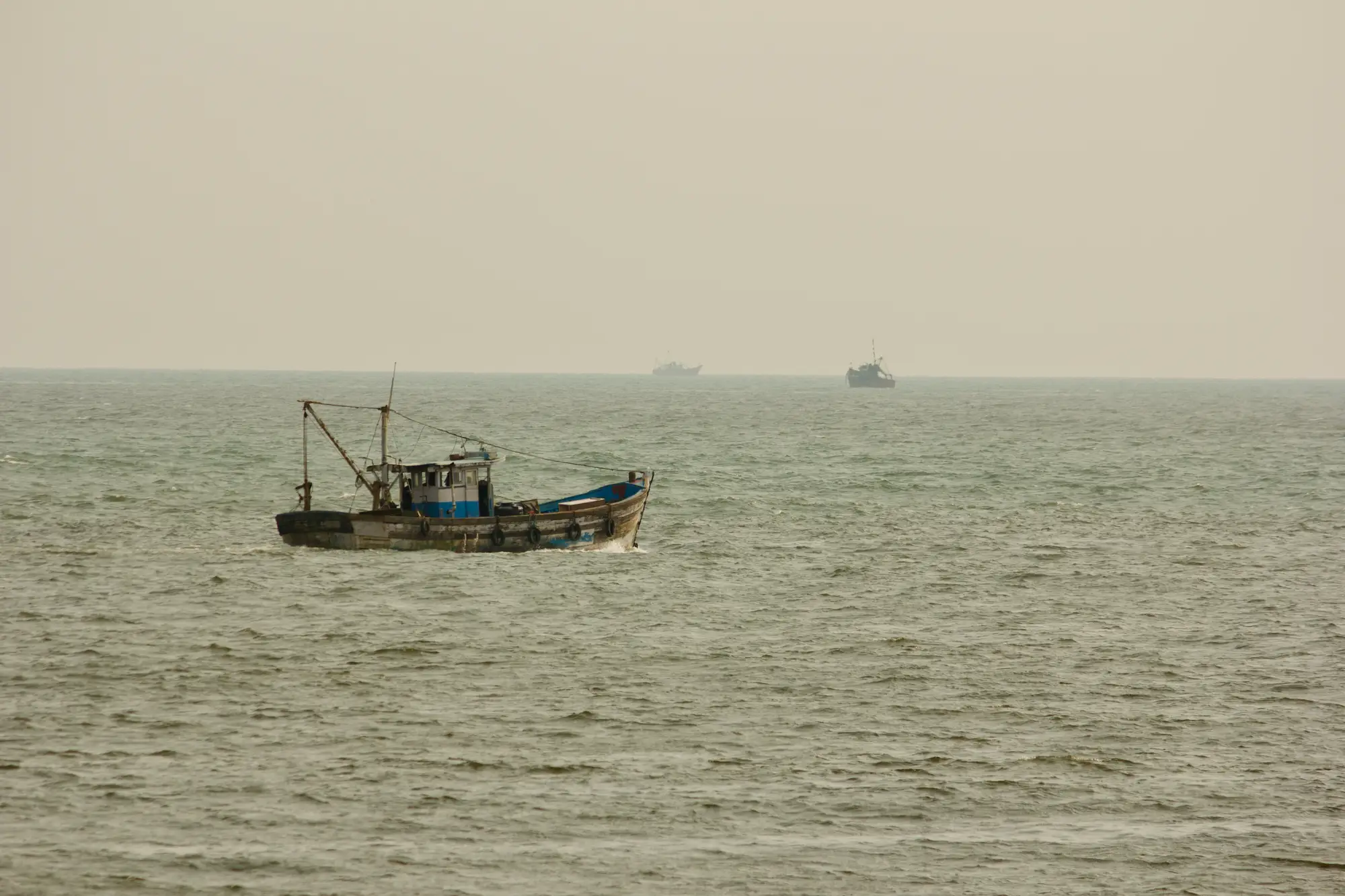 A fishing boat working a hazy, flat sea with two more vessels on the horizon